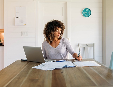 IMAGE: Woman at table with laptop making notes.