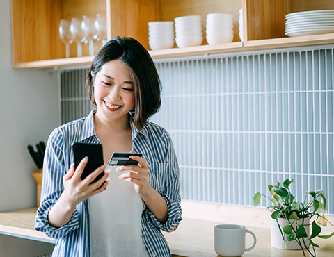 IMAGE: woman in kitchen holding credit card looking at phone and smiling.