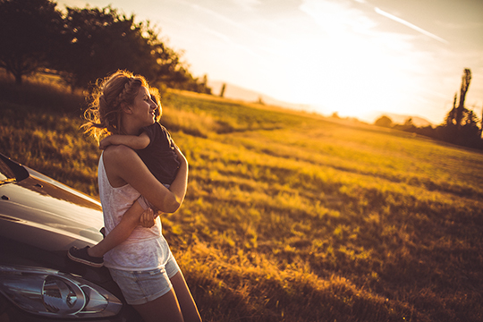 IMAGE: Woman holding small child and leaning against hood of car with sunny field in background