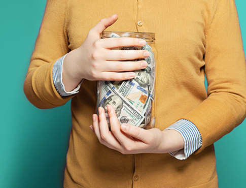 IMAGE: Close crop of midsection, woman in gold cardigan holding glass jar filled with paper currency.