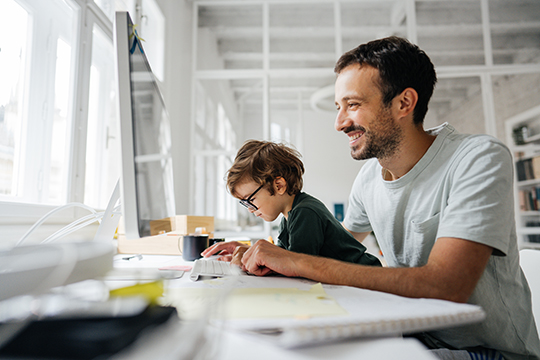 IMAGE: Man sitting at computer desk smiling with child working beside him