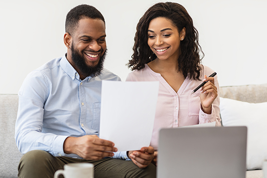 IMAGE: Couple sitting together on a couch and looking at a piece of paper and laptop smiling