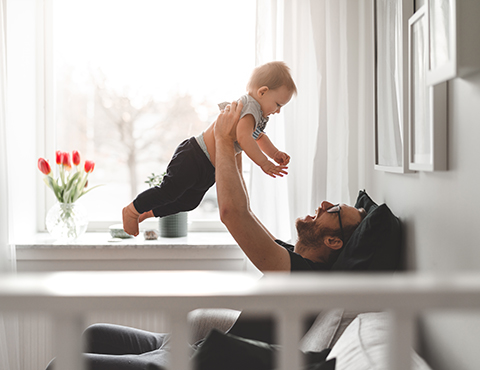 IMAGE: Man sitting in chair holding toddler above him and laughing.