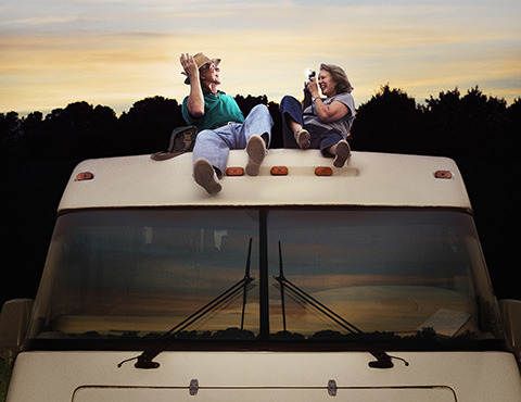 IMAGE: Mature couple reclined on roof of RV at sunset. Man posing for photo, woman with camera in-hand.