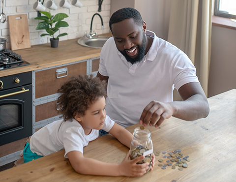 IMAGE: man sitting at counter with young son putting coins into glass jar.