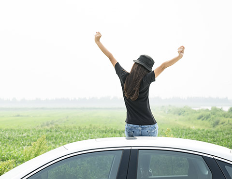 IMAGE: Girl facing away from camera, sitting on car with arms stretched high in air.