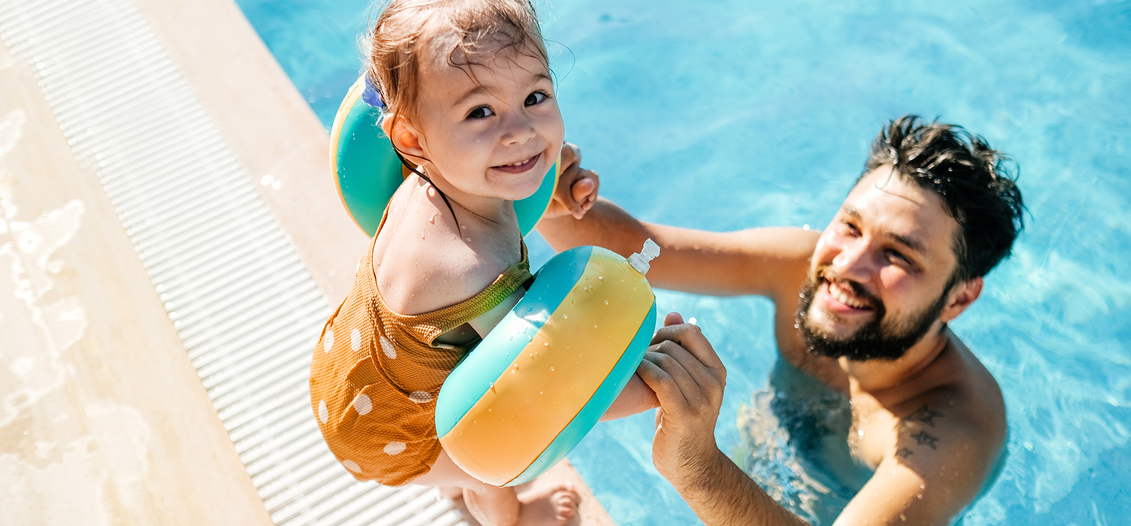 IMAGE: Man in a pool holding hand of young daughter standing at the edge and looking back at the camera