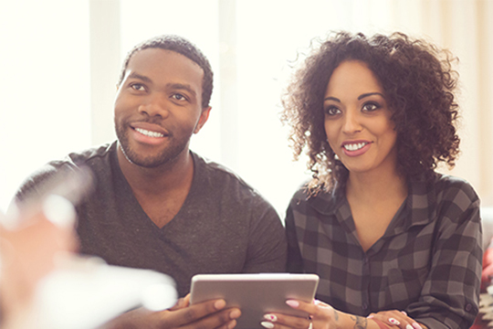 IMAGE: Couple holding tablet together and smiling at insurance agent