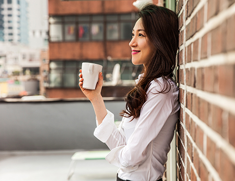 IMAGE: Woman standing against brick wall with cup of coffee looking happy.