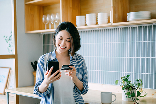 IMAGE: woman in kitchen holding credit card looking at phone and smiling.