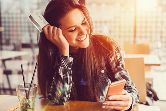 IMAGE: Young woman smiling and holding phone and debit card inside cafe