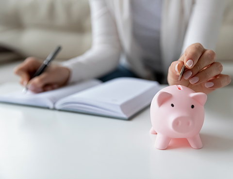 IMAGE: woman sitting with notepad open and putting a coin into a piggy bank