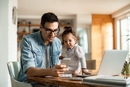 IMAGE: Man looking at phone and laptop while little girl sits on the table next to him looking at phone.
