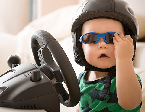 IMAGE: Toddler with helmet and sunglasses sitting at toy steering wheel.