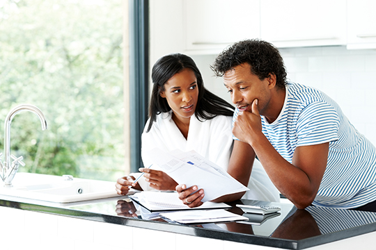 IMAGE: Couple sitting at kitchen counter looking worried at paperwork